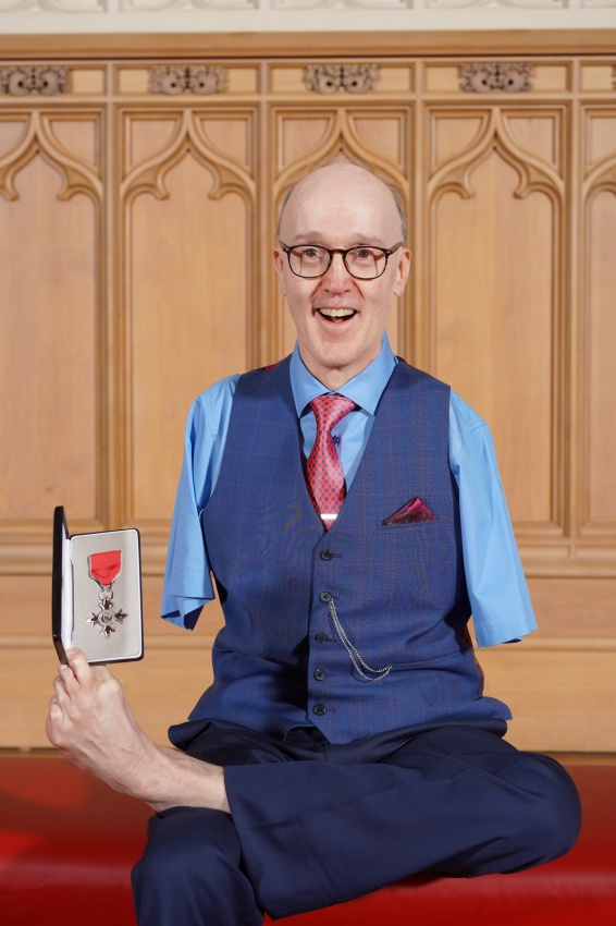 Brian Gault holding up his MBE medal in it's case with his foot