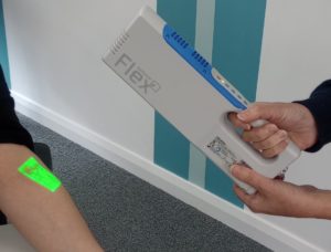 A person's hands holding a vein viewer over another person's arm, with a green light patch on that arm showing where veins are
